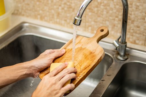 A person gently washing a wooden cutting board with a sponge under running water, showing proper care for the healthiest cutting boards.