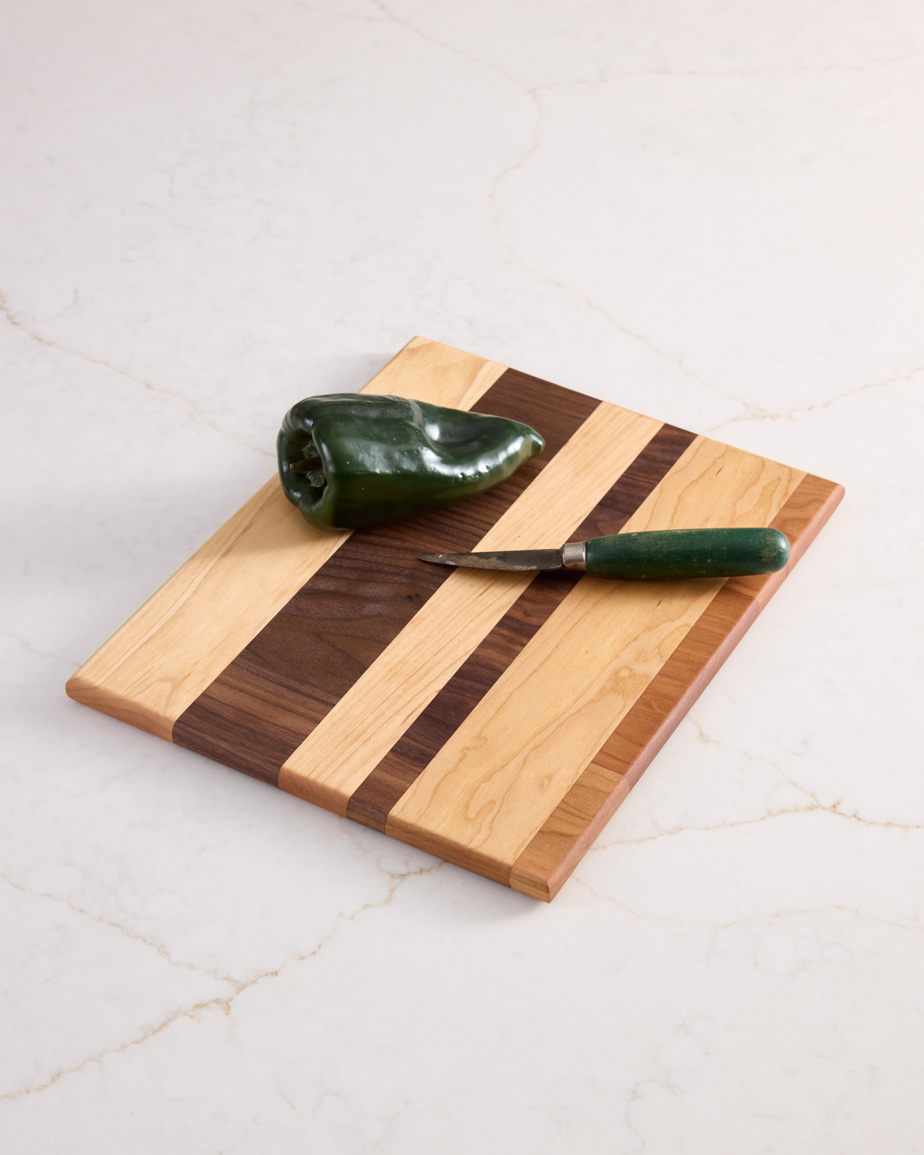 Tri-wood cutting board with jalapeño and green knife, made from walnut, maple, and cherry, styled on white marble.