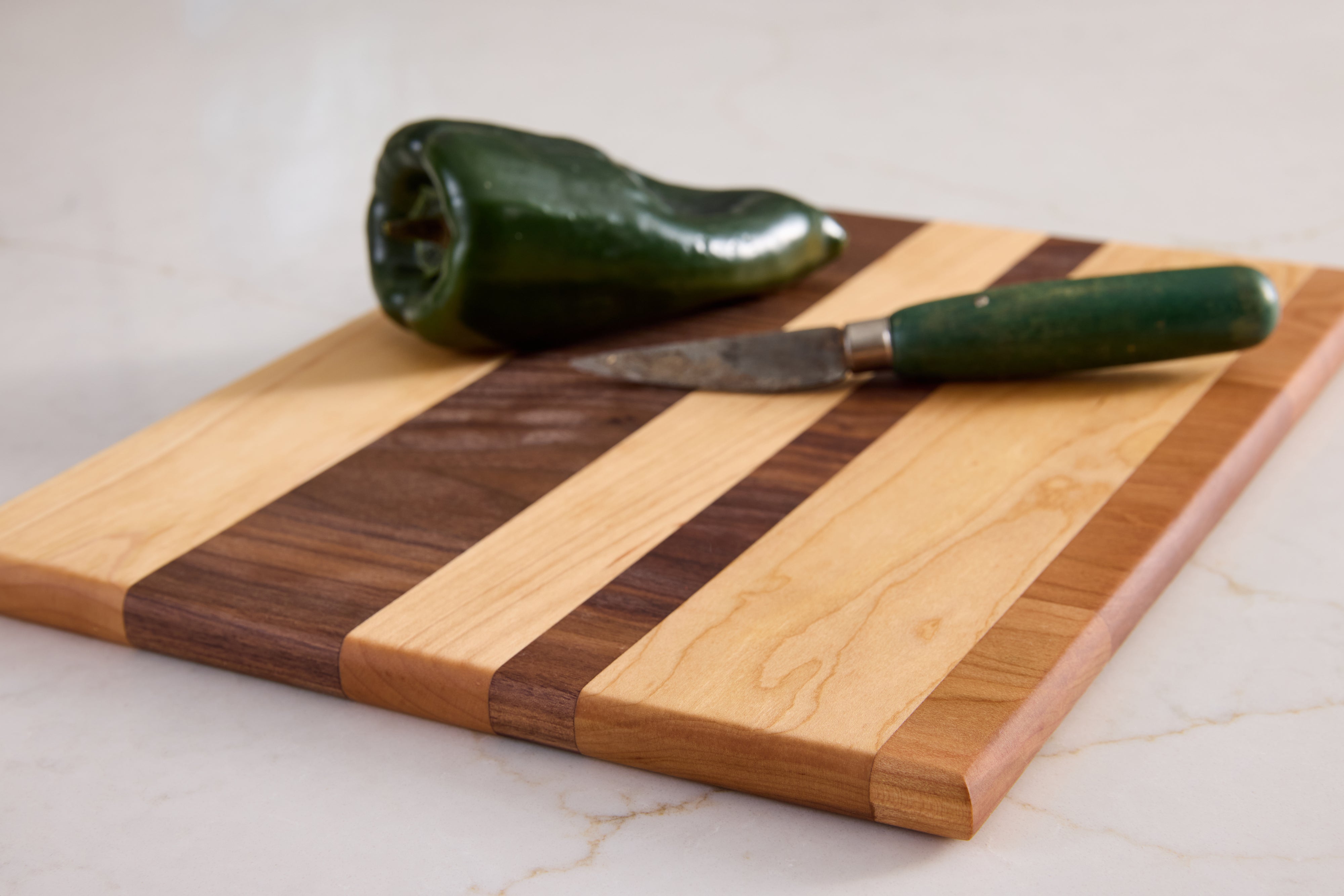 Walnut, cherry, and maple cutting board with green knife and jalapeño, showcasing prep-ready striped wood grain on marble.