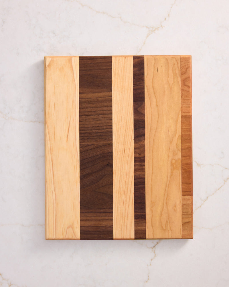 Small striped wooden cutting board made from cherry, walnut, and maple, slanted on marble, ideal for modern wood tray decor.
