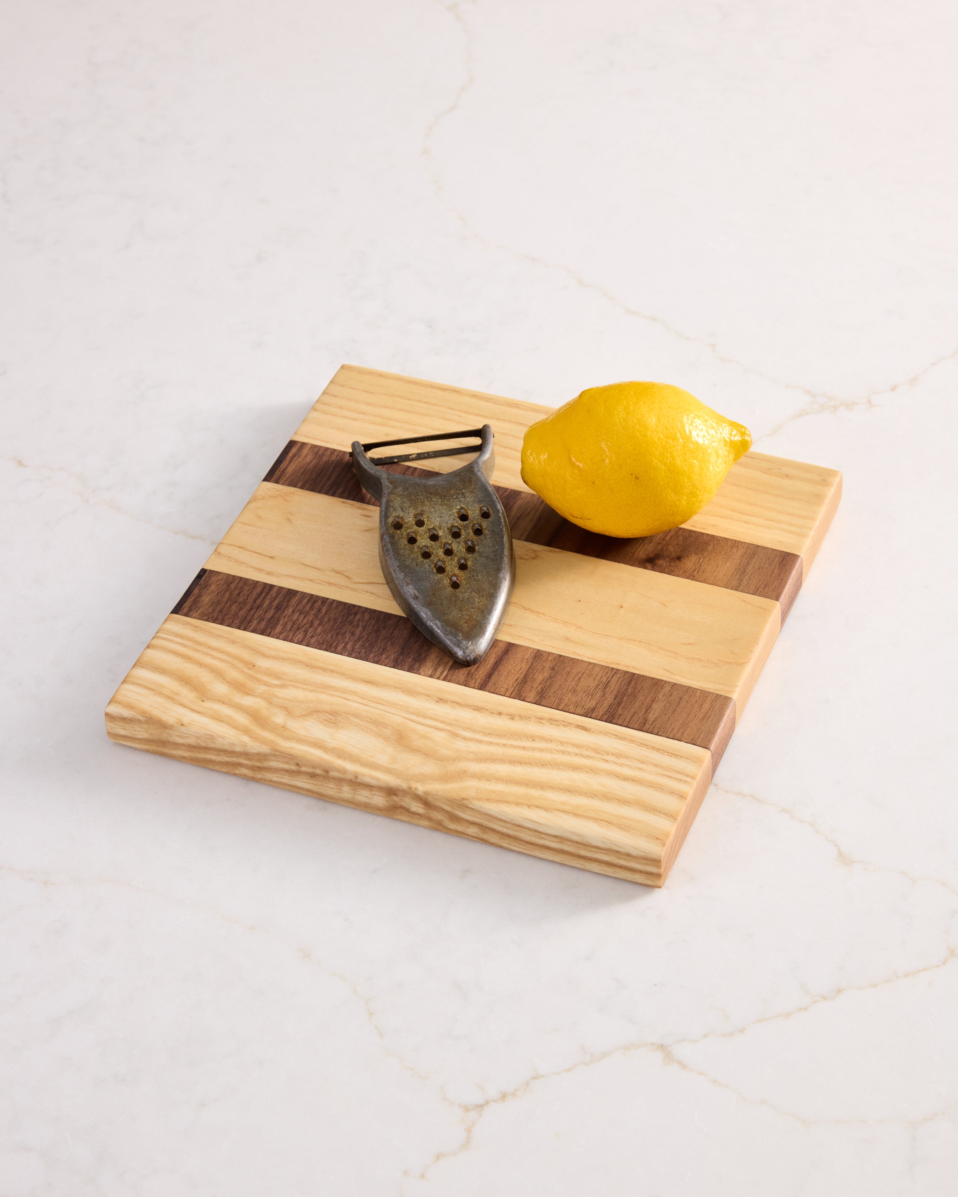 Minimalist wooden garnish board made of maple and walnut, shown with lemon and peeler on a clean white marble surface.