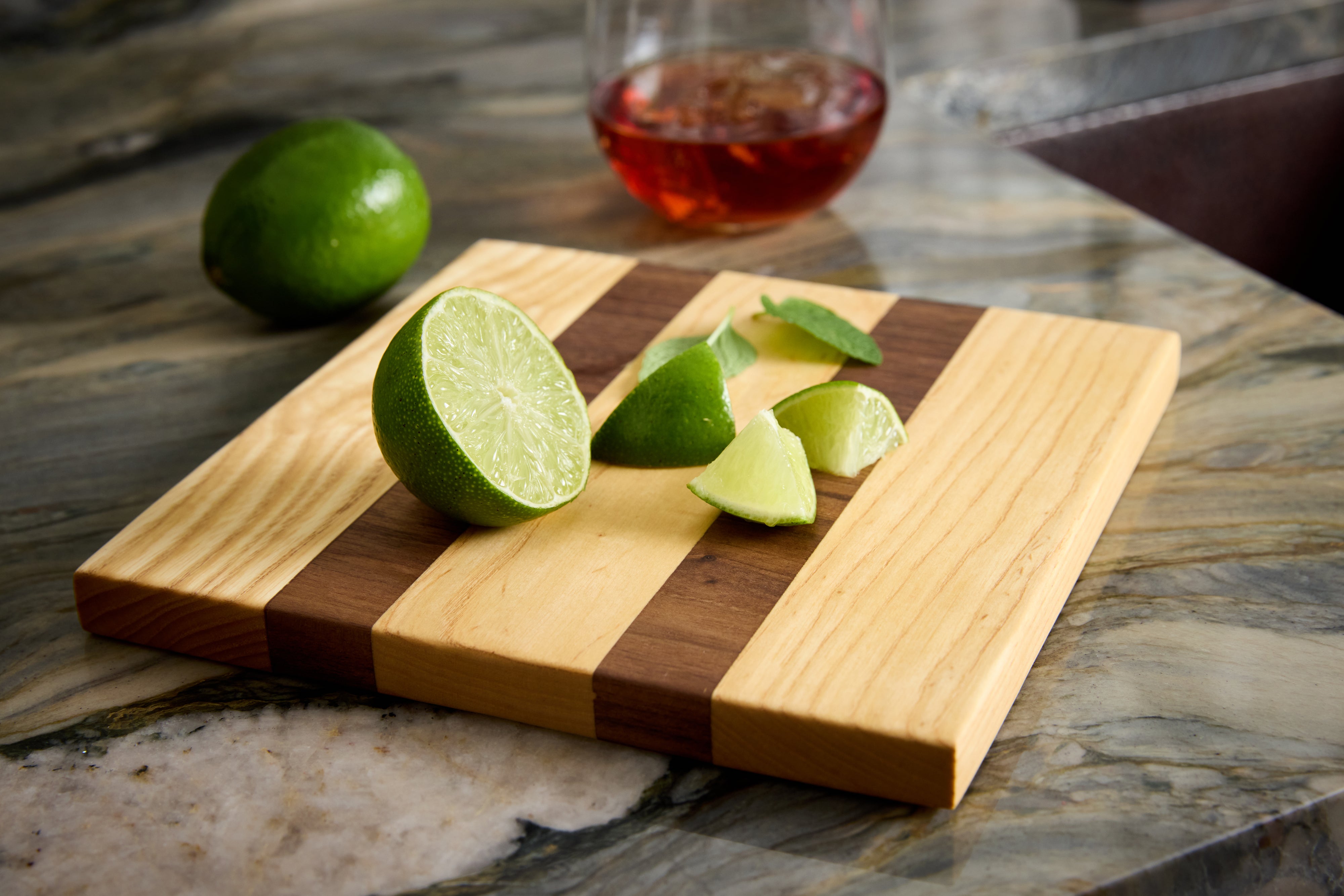 Maple and walnut garnish board styled with lime slices, mint leaves, and cocktail — a non-toxic prep board for home bars.