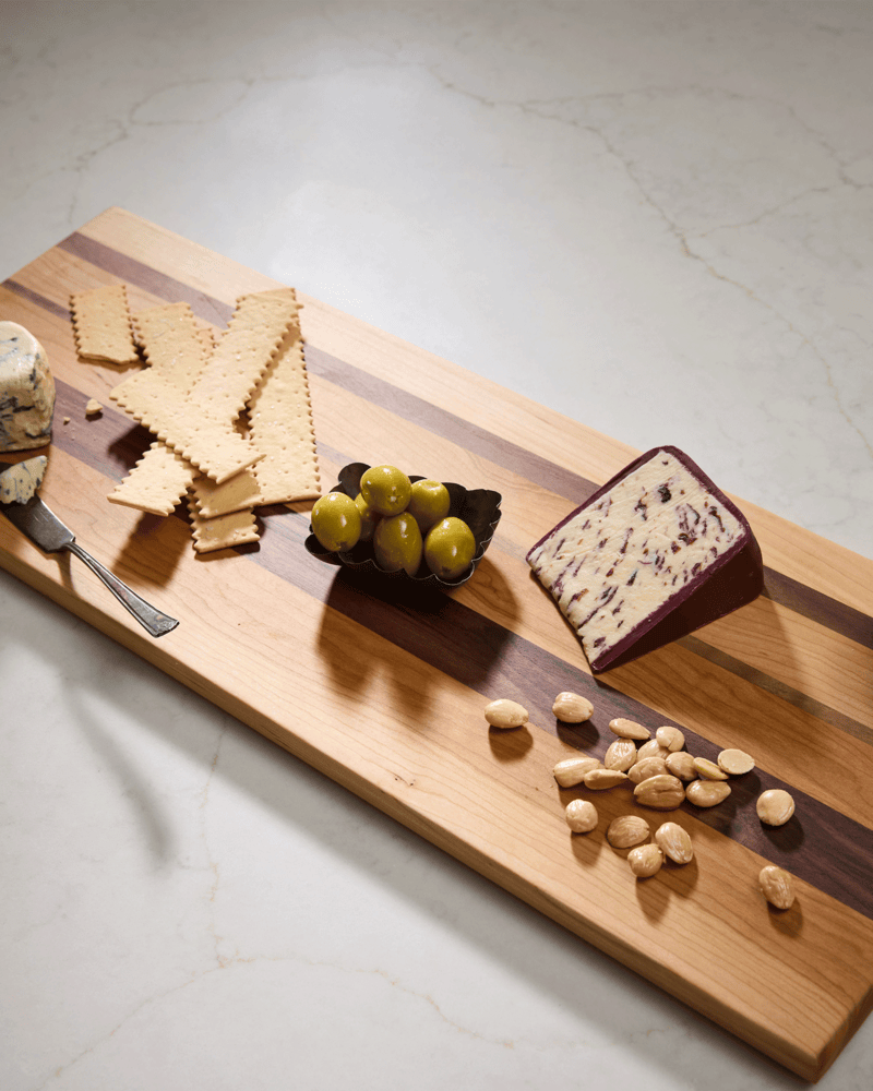 Tri-wood maple, walnut, and cherry cutting board with cheese, olives, nuts, and honey, styled with flowers on white marble.