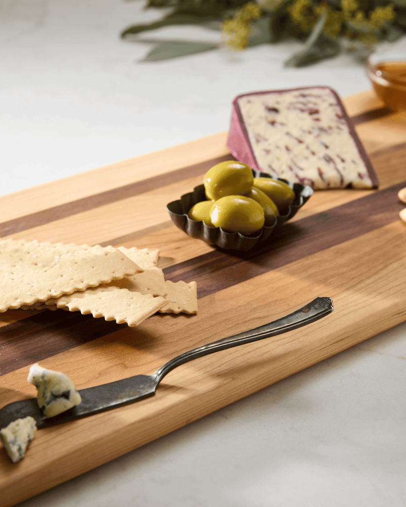 Slanted walnut and maple charcuterie cutting board styled with marbled cheese, crackers, olives, honey, and nuts.