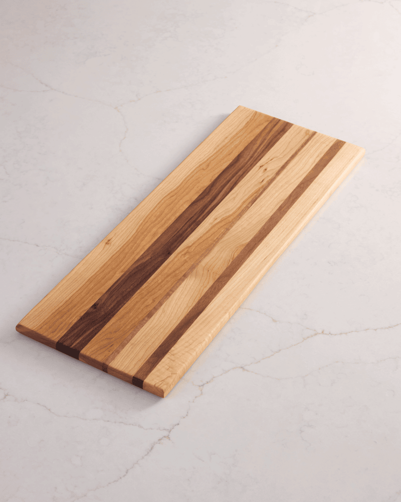 Tri-wood walnut, cherry, and maple cutting board with striped grain pattern, angled on a white marble kitchen surface.