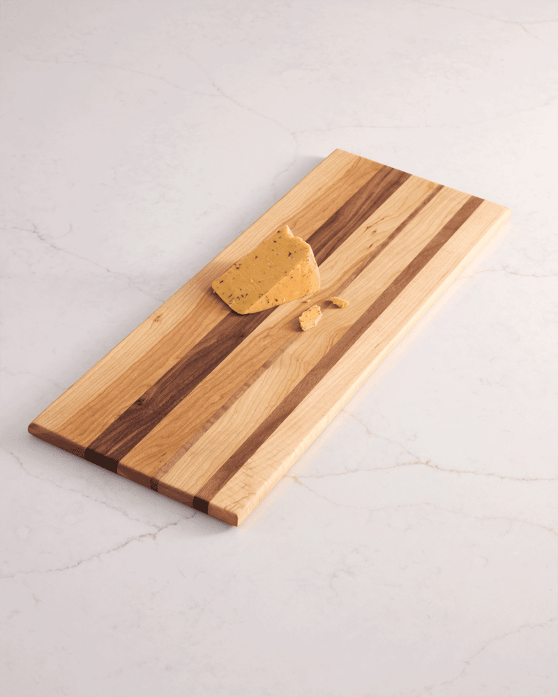 A tri-wood cherry, maple, and walnut cutting board with a block of cheese on a white marble surface- best cutting board.