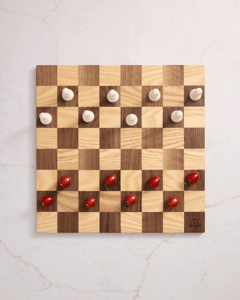 Handmade walnut and maple chess board styled with cherry tomatoes and chess pieces on white marble — unique board decor.